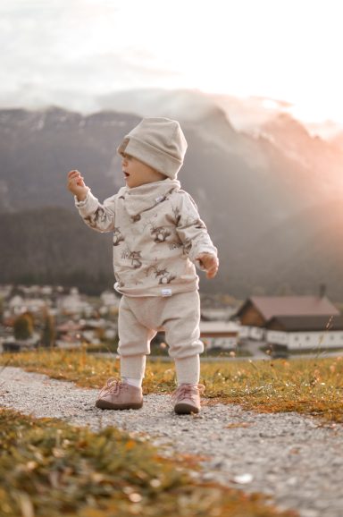 Kleines Kind in warmer Kleidung, steht auf einem Weg mit Bergen im Hintergrund.