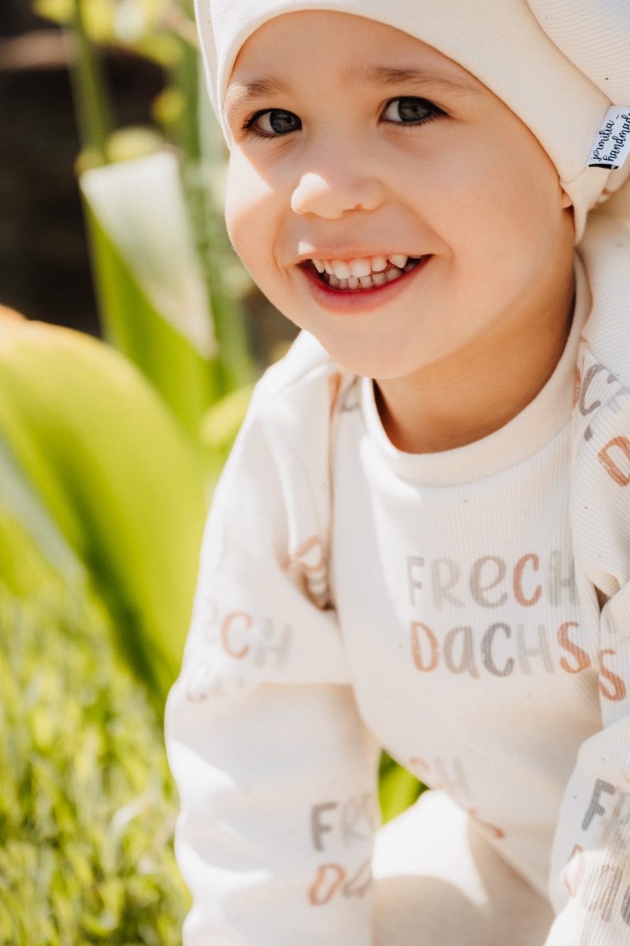 Kleines Kind in hellem Outfit sitzt auf einer Steinmauer, umgeben von Gras und Blumen.