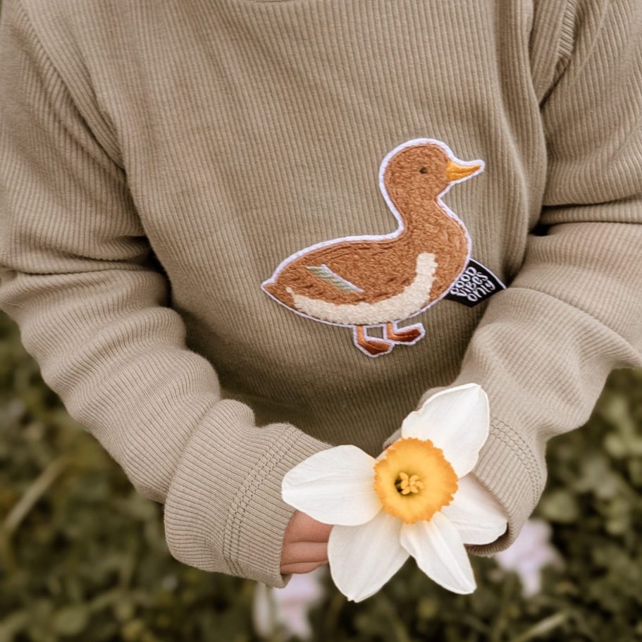 Ein Kind hält eine weiße Blume mit einer gelben Mitte und trägt ein braunes Shirt mit Entenmotiv.